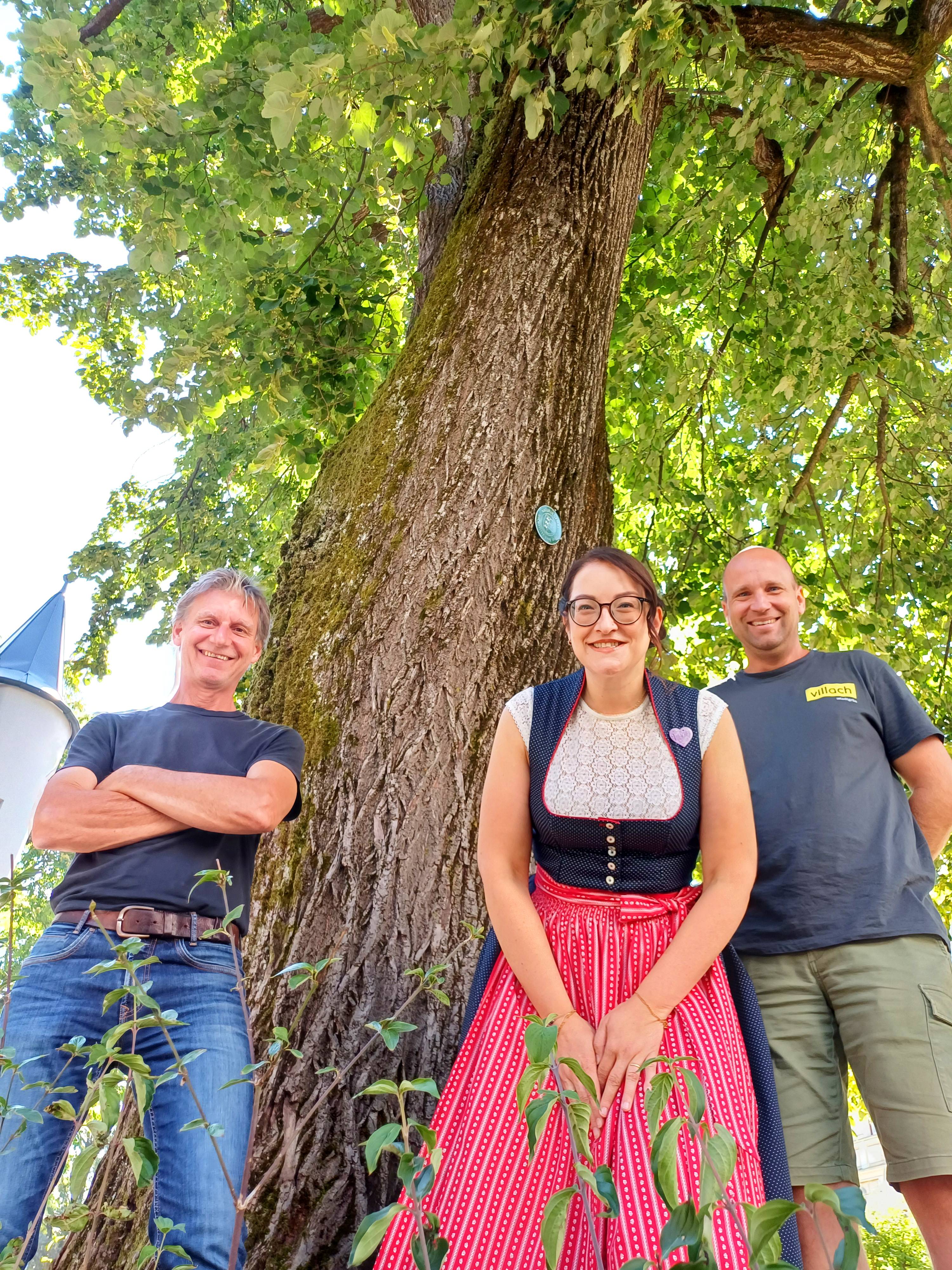 Drei Personen stehen vor einem Baum, eine Frau im Dirndl, ein Mann in Jeans und ein Mann mit T-Shirt.