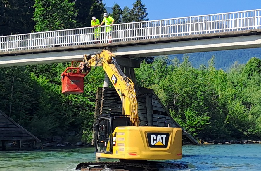 Bagger im Fluss hebt roten Greifer unter Brücke mit zwei Arbeitern an