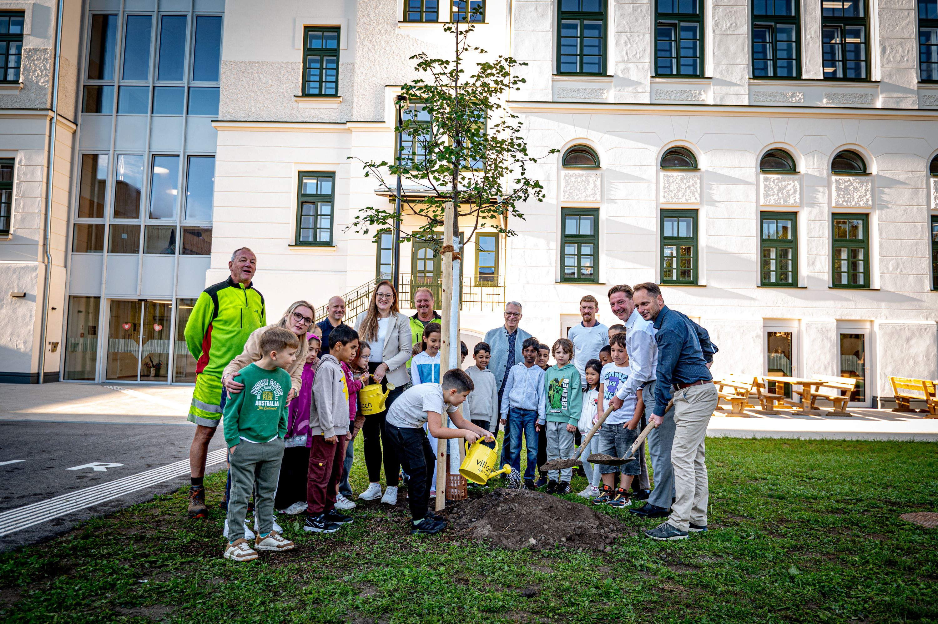 Schüler und Lehrer pflanzen mit Schaufeln und Gießkanne einen Baum vor dem Schulgebäude in Wien.