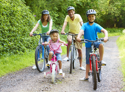 Fröhliche Familie beim Fahrradausflug. Zwei Erwachsene, zwei Kinder, alle tragen unterschiedlich färbige T-Shirts und tragen einen Fahrradhelm.