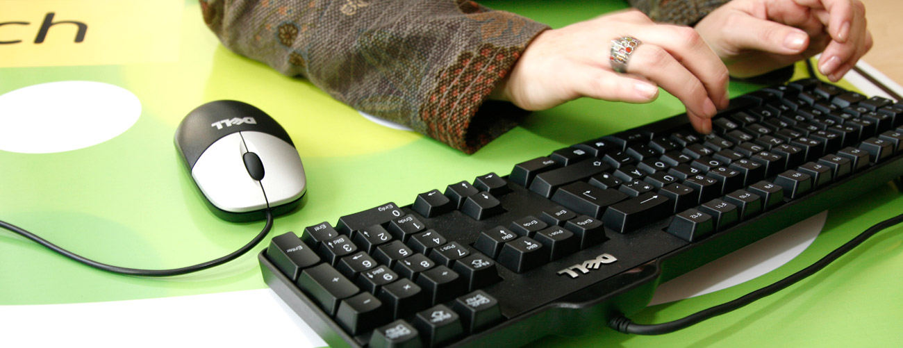 Gepflegte Frau tippt auf Computertastatur, eine Computer-Maus befindet sich daneben, im Hintergrund sieht man einen Teil des Logos der Stadt Villach