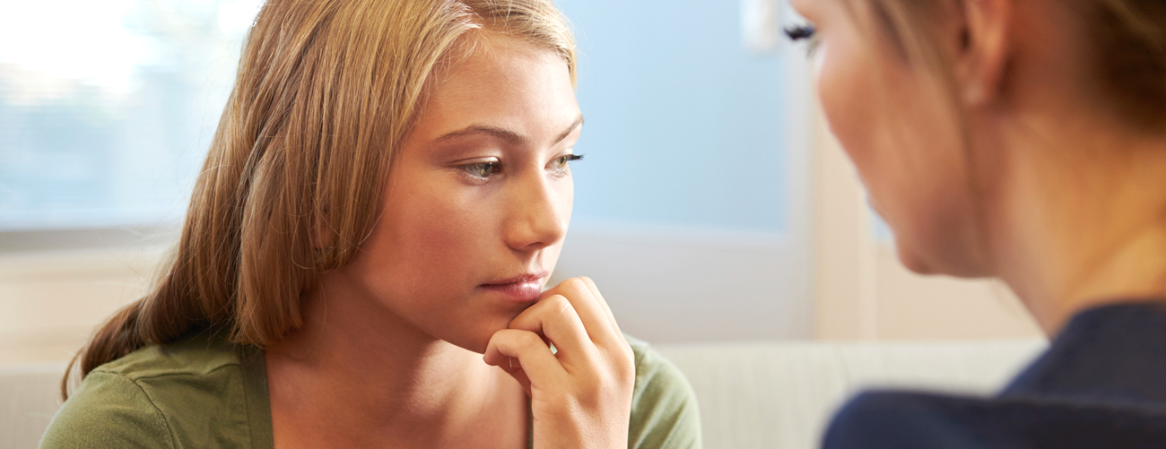 Junges Mädchen blickt ernst zur Seite. Vor ihr sitzt eine Frau die zu ihr blickt und ihr helfen will.