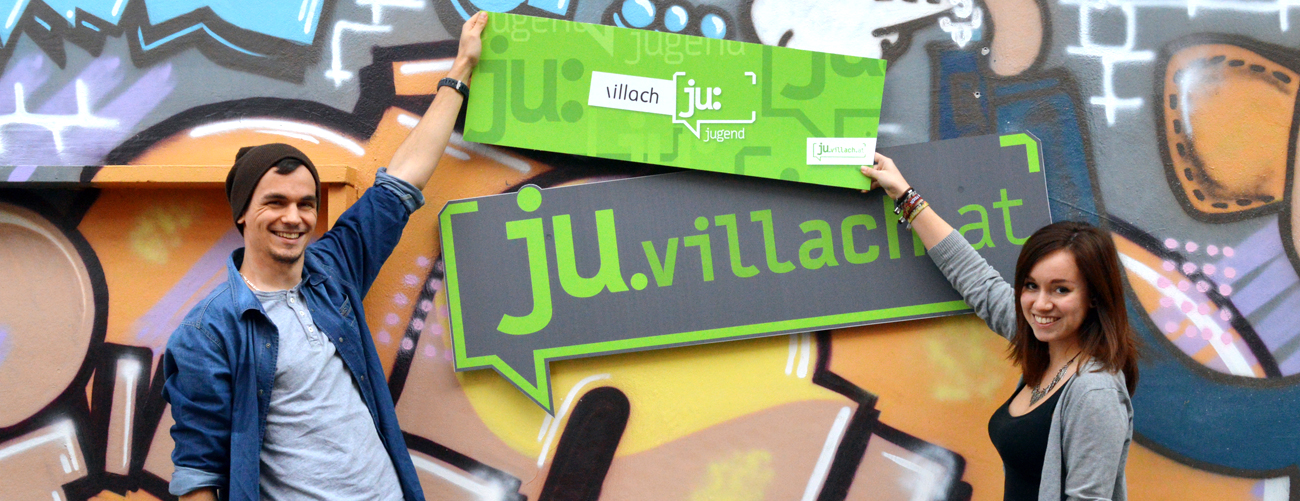 Zwei Jugendliche (Mädchen und Bub) halten ein großes Schild (das Jugend-Logo der Stadt Villach) auf die mit Graviti besprühten Fassade unseres Jugendzentrums in der Gerbergasse in Villach.