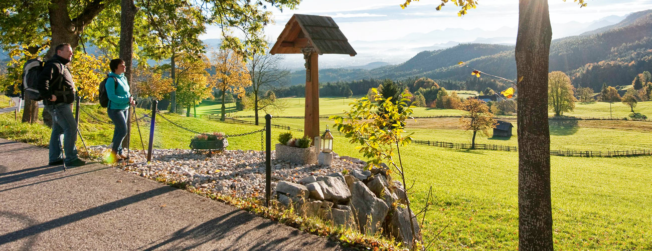 Pilgern in Kärnten - 2 Wanderer mit Stöcken die an einem Wanderweg entlang bei einem Materl vorbeigehen. Es ist später Nachmittag und die Sonne steht tief.